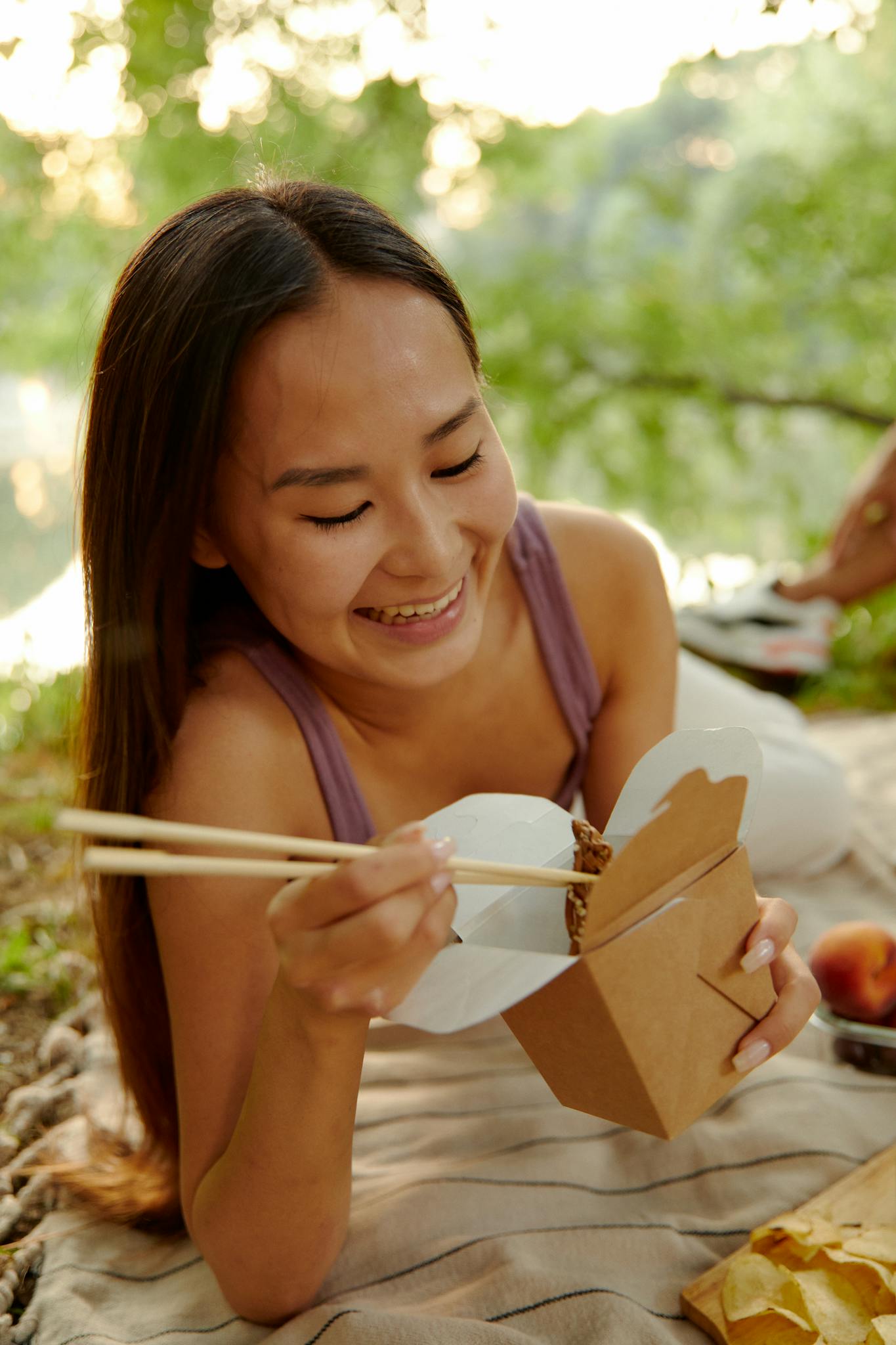 Asian woman smiling while enjoying a picnic outdoors with takeout food and chopsticks on a sunny day.