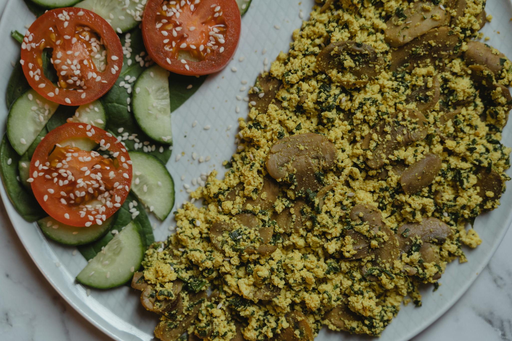 Healthy vegan meal with tofu scramble, cucumbers, and tomatoes on a marble surface.