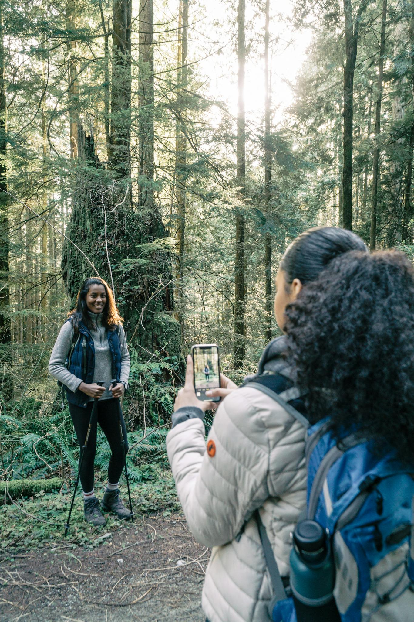 Two women hiking in a forest, one taking a photo of the other with a smartphone.
